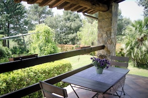 A wooden bench and table on a patio with a stone wall, surrounded by lush greenery and trees.