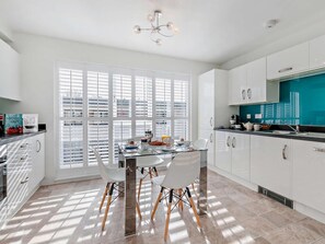 A modern kitchen with white cabinetry, a dining table with chairs, and a large window with blinds. - 3 Bed in St. Austell (oc-65572) (Charlestown)