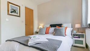 Ein ordentlich bezogenes Bett mit Kissen, ein Nachttisch mit Lampe, Bücher auf einem Beistelltisch und ein gerahmtes Bild an der Wand.