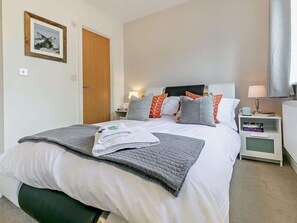 A neatly made bed with pillows, a bedside table with a lamp, a nightstand with books, and a framed picture on the wall. - 3 Bed in St. Austell (oc-65572) (Charlestown)