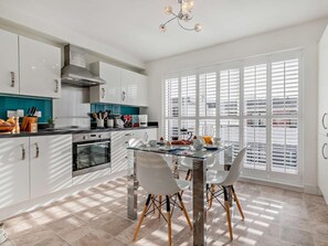 A modern kitchen with white cabinets, a stainless steel range hood, and a dining area with a glass table and white chairs. - 3 Bed in St. Austell (oc-65572) (Charlestown)