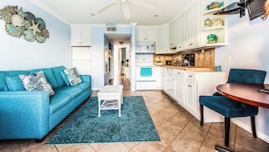A modern kitchen with white cabinets, a microwave, and a coffee maker. There is a teal sofa with a matching ottoman and a blue chair. - True Beachfront Condo - Steps to Sand! No Seawall Crossing. 7.7 miles from Port. (Galveston)