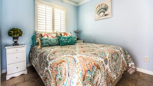 A bedroom with a bed, a nightstand, a window with blinds, a wall-mounted fan, and a small potted plant. - True Beachfront Condo - Steps to Sand! No Seawall Crossing. 7.7 miles from Port. (Galveston)