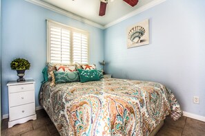Une chambre à coucher comprenant un lit, une table de chevet, une fenêtre avec des stores, un ventilateur fixé au mur et une petite plante en pot.