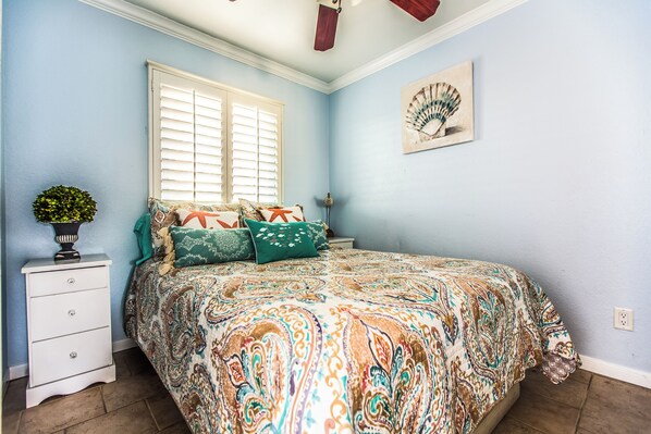 A bedroom with a bed, a nightstand, a window with blinds, a wall-mounted fan, and a small potted plant. - True Beachfront Condo - Steps to Sand! No Seawall Crossing. (Galveston)