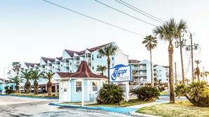 A resort with multiple blue buildings, a small kiosk, and palm trees. - True Beachfront Condo - Steps to Sand! No Seawall Crossing. 7.7 miles from Port. (Galveston)