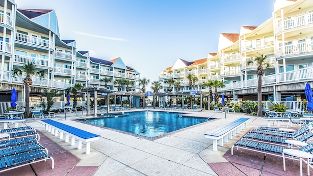 A hotel pool area with multiple lounge chairs and benches, surrounded by multi-story buildings with balconies. - True Beachfront Condo - Steps to Sand! No Seawall Crossing. 7.7 miles from Port. (Galveston)