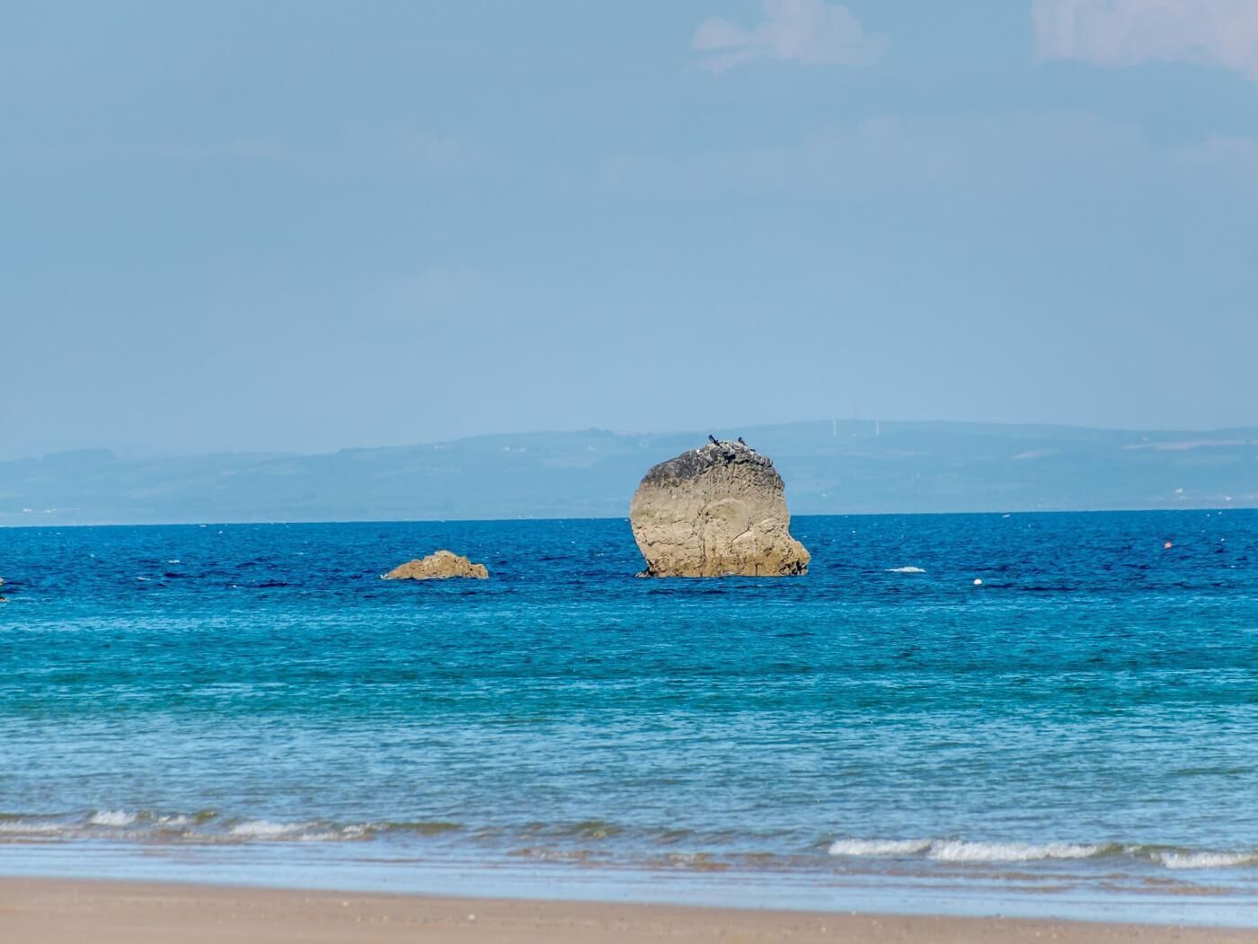 A beach with clear blue water, a large rock formation, and distant mountains.