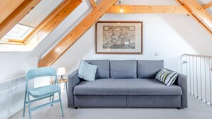 Un dormitori tipus loft amb sostre inclinat, un sofà gris, una cadira blava, una obra d'art emmarcada i un òcul de bou.