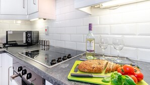 Una cuina moderna amb armaris blancs, un microones integrat, una cuina a gas, una taula de tall amb pa i tomàquets, i una ampolla de vi.