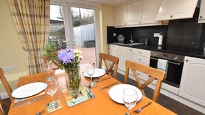 Un comedor con una mesa de madera dispuesta para cuatro personas, un florero con flores y una cocina con armarios blancos y encimera negra.