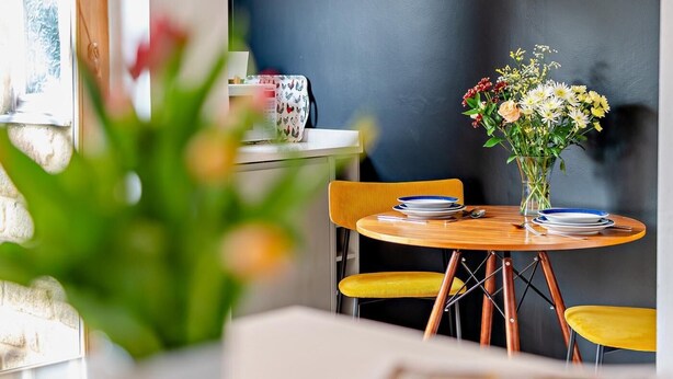 Un'area pranzo con un tavolo rotondo, sedie gialle, un vaso con fiori e una lampada a sospensione in rame.