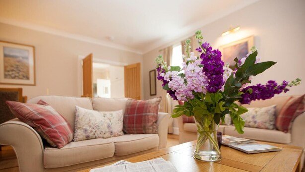 Ein Wohnzimmer mit einem beigen Sofa, einem Couchtisch mit einer Zeitung und einer Vase mit violetten Blumen.
