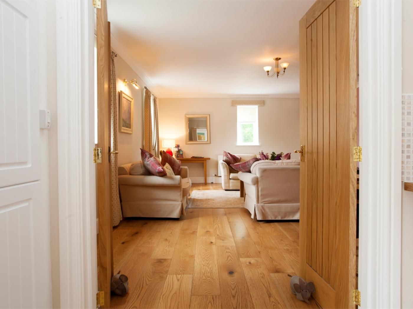 A hallway leading to a living area with two sofas and a table, wooden flooring, and a chandelier.