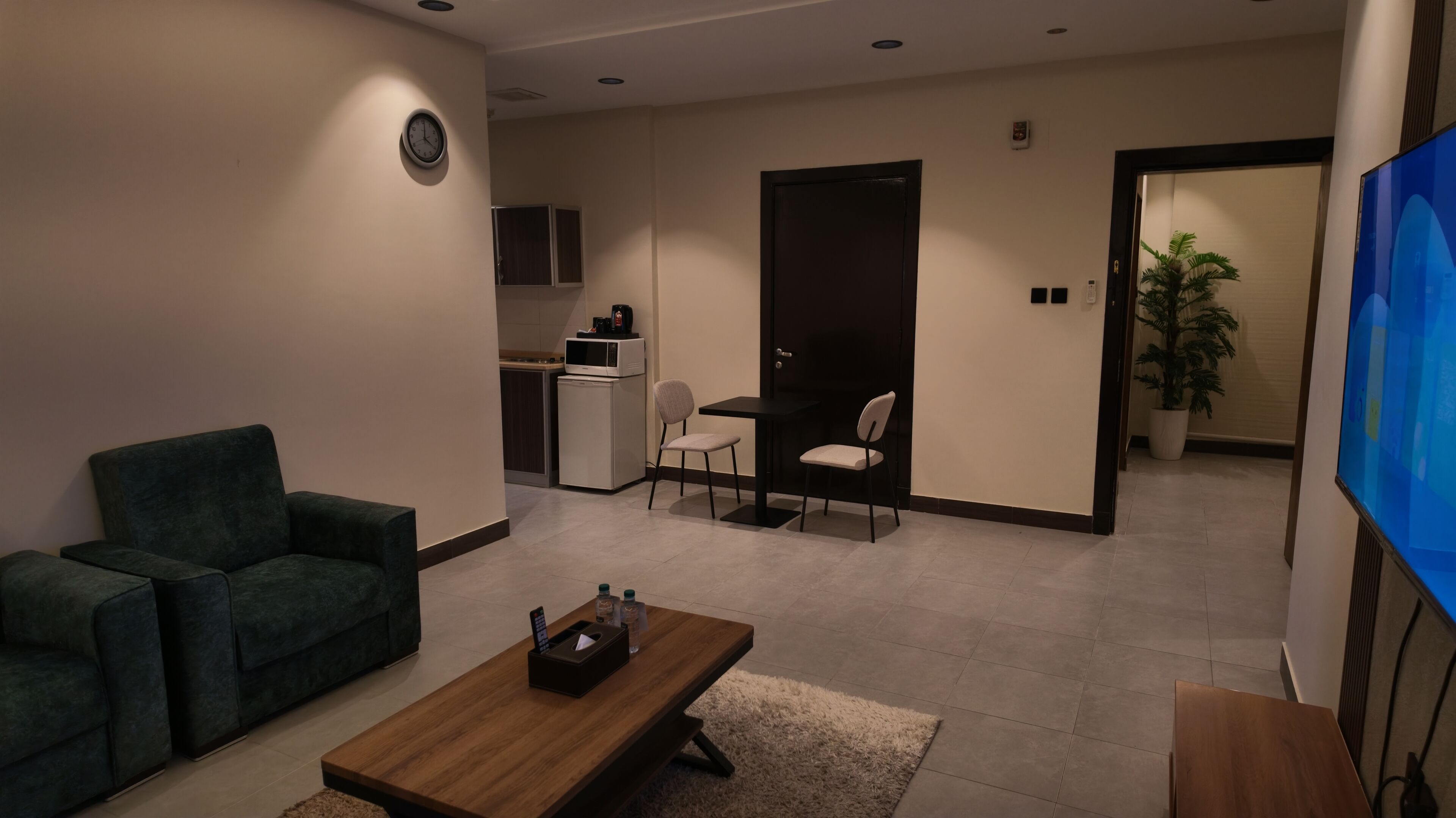 A modern living room with a sofa, a coffee table, and a TV. There is a kitchen area with a microwave and a refrigerator.