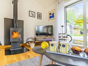 A cozy living room with a wood-burning stove, a TV, a dining table with mugs, and a view of the outdoors. - 3 Bed in Nr Masham (oc-g0149) (Healey)