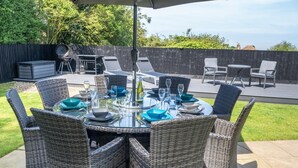An outdoor dining area with wicker chairs, a glass table, and a barbecue grill in the background. - 4 Bed in Cromer (oc-82588) (Cromer)