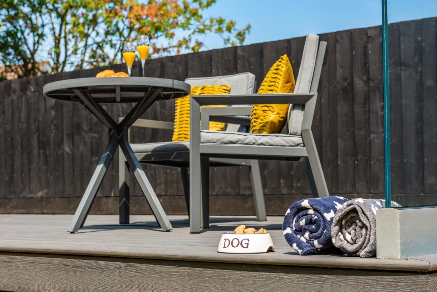 Un espace extérieur aménagé avec une table ronde, une chaise avec des coussins, une couverture pliée et un bol pour chien.