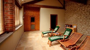 Ein Wintergarten mit Holzbalken, einer Steinwand und einer Backstein-Säule. Es befinden sich Lounge-Stühle mit grünen Kissen darin.