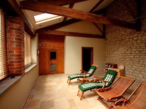 Ein Wintergarten mit Holzbalken, einer Steinwand und einer Backstein-Säule. Es befinden sich Lounge-Stühle mit grünen Kissen darin.