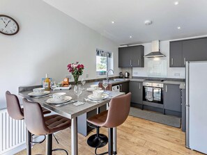 A modern kitchen with a dining area, featuring a clock, a vase with flowers, and a window with a view. - 2 Bed in Camborne (oc-82635) (Camborne)