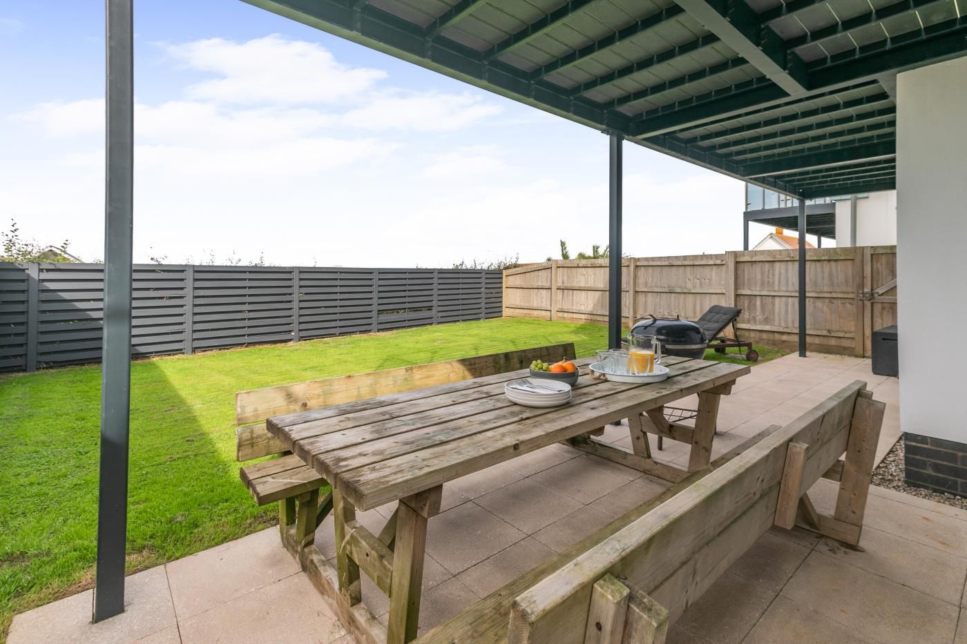 A wooden picnic table with a beverage tray and a grill in a backyard with a fenced lawn.