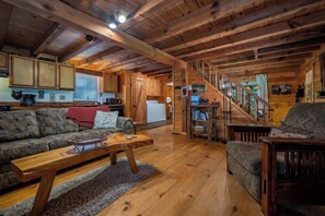 A log cabin interior with a wooden staircase, a sofa, a coffee table, and a kitchen area.