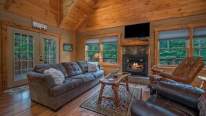 A wooden cabin interior with a fireplace, leather sofas, a TV, and large windows. - Private LogHome/ HotTub/Lake Lure/pet (Lake Lure)