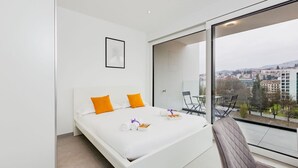 Ein modernes Hotelzimmer mit einem großen Bett, Blick auf die Stadt und einem Sitzgelegenheit am Balkon.