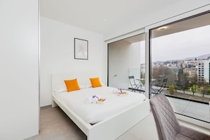 Ett modernt hotellrum med en stor säng, utsikt över staden och en balkong med sittplatser.