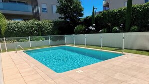Uma piscina retangular com deck ladrilhado, cercada por uma cerca branca e uma sebe ao fundo.