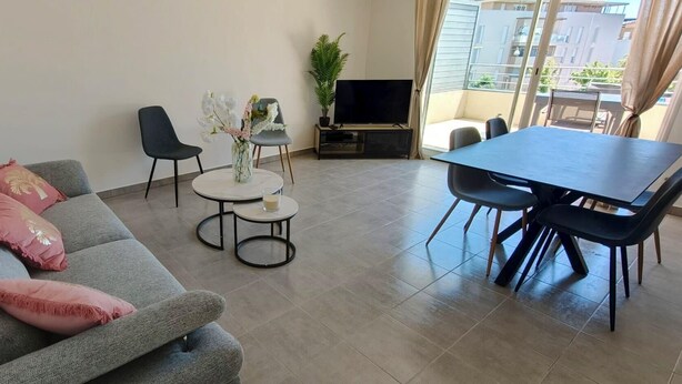 Uma sala de estar com um sofá cinzento, duas mesinhas redondas, uma televisão e vista para uma varanda.