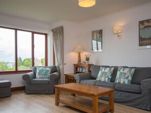 A living room with a sofa, armchair, and coffee table, a bookshelf, and a view through large windows. - 4 Bed in Swanage (oc-dc020) (Studland)