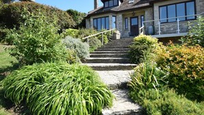 Una scala in pietra conduce a un edificio in pietra con ampie finestre e un giardino con diverse piante.