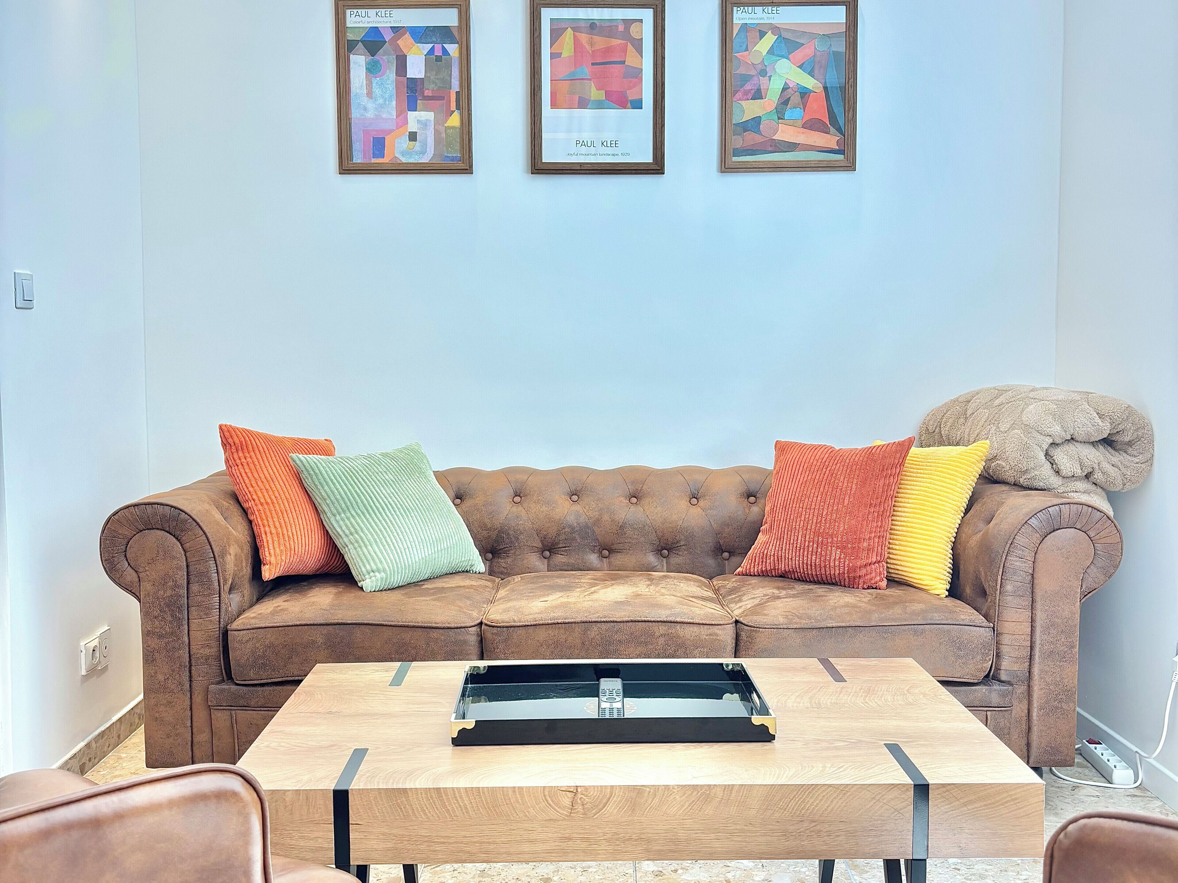 Un canapé en cuir marron agrémenté de coussins colorés, une table basse en bois avec un plateau, et trois œuvres d’art abstraites encadrées accrochées au mur.