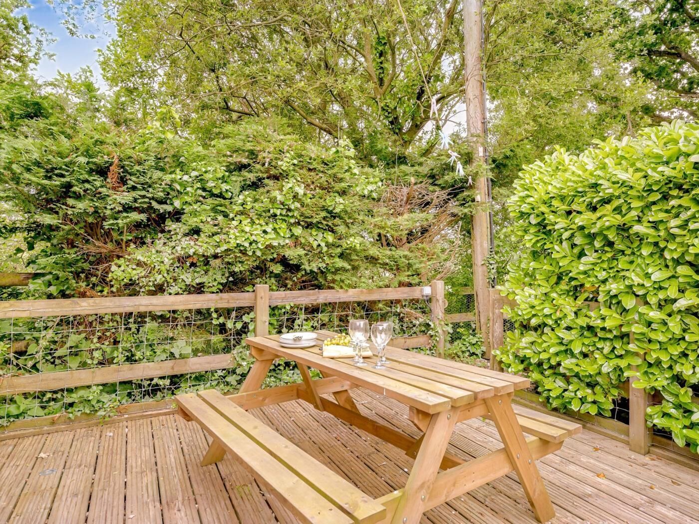 Un terrazzo in legno con tavolo da picnic, circondato da vegetazione lussureggiante e un recinto di legno.