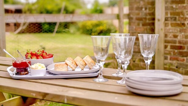 A picnic setup with food and drinks on a wooden table, set against a brick wall and greenery. - 3 Bed in Wareham (oc-dc157) (Stoborough)