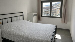 Een slaapkamer met een bed, een kaptafel, een raam met gordijnen en uitzicht op gebouwen.