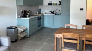 Een moderne keuken met blauwgroene kastjes, een eettafel met houten stoelen en een grote witte hanglamp.