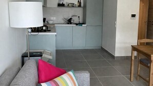 Een moderne woonkamer met een grijze bank, een kleurrijk gestreept kussen, een witte lamp en een houten eettafel.