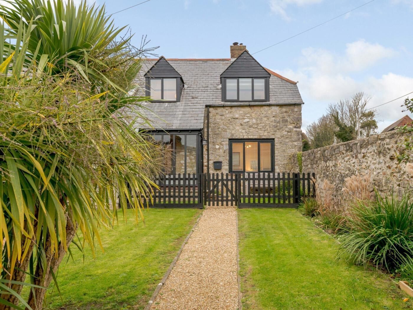 A stone house with a gravel path leading to the entrance, surrounded by greenery and a stone wall.