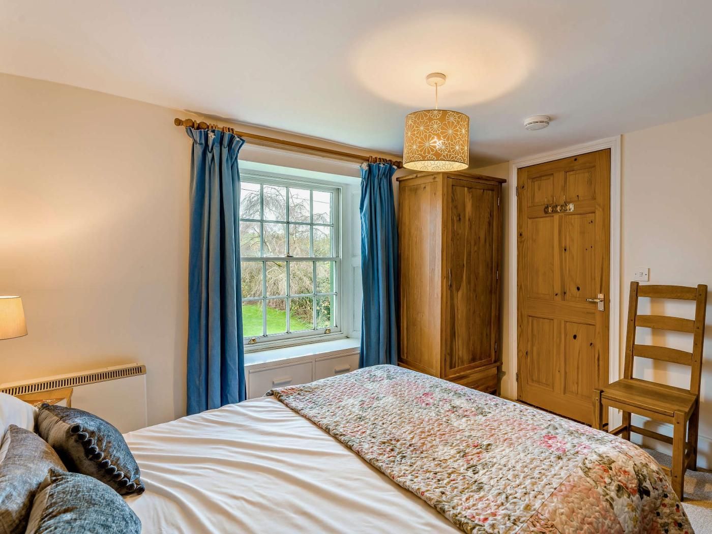 A bedroom with a bed, a window with curtains, a wooden wardrobe, and a chair.