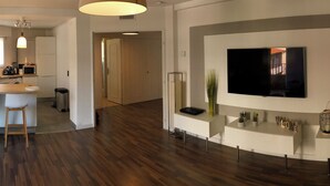 Uma sala de estar moderna com televisão de tela plana, piso de madeira, área de cozinha com micro-ondas e uma janela grande.