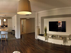 A modern living room with a flat-screen TV, a wooden floor, a kitchen area with a microwave, and a large window. - Bright apartment near center and beaches (Cannes)