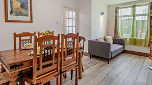 A dining area with a wooden table and chairs, a grey sofa, a window with curtains, and a framed painting on the wall. - 2 Bed in Mablethorpe (oc-74809) (Mablethorpe and Sutton)