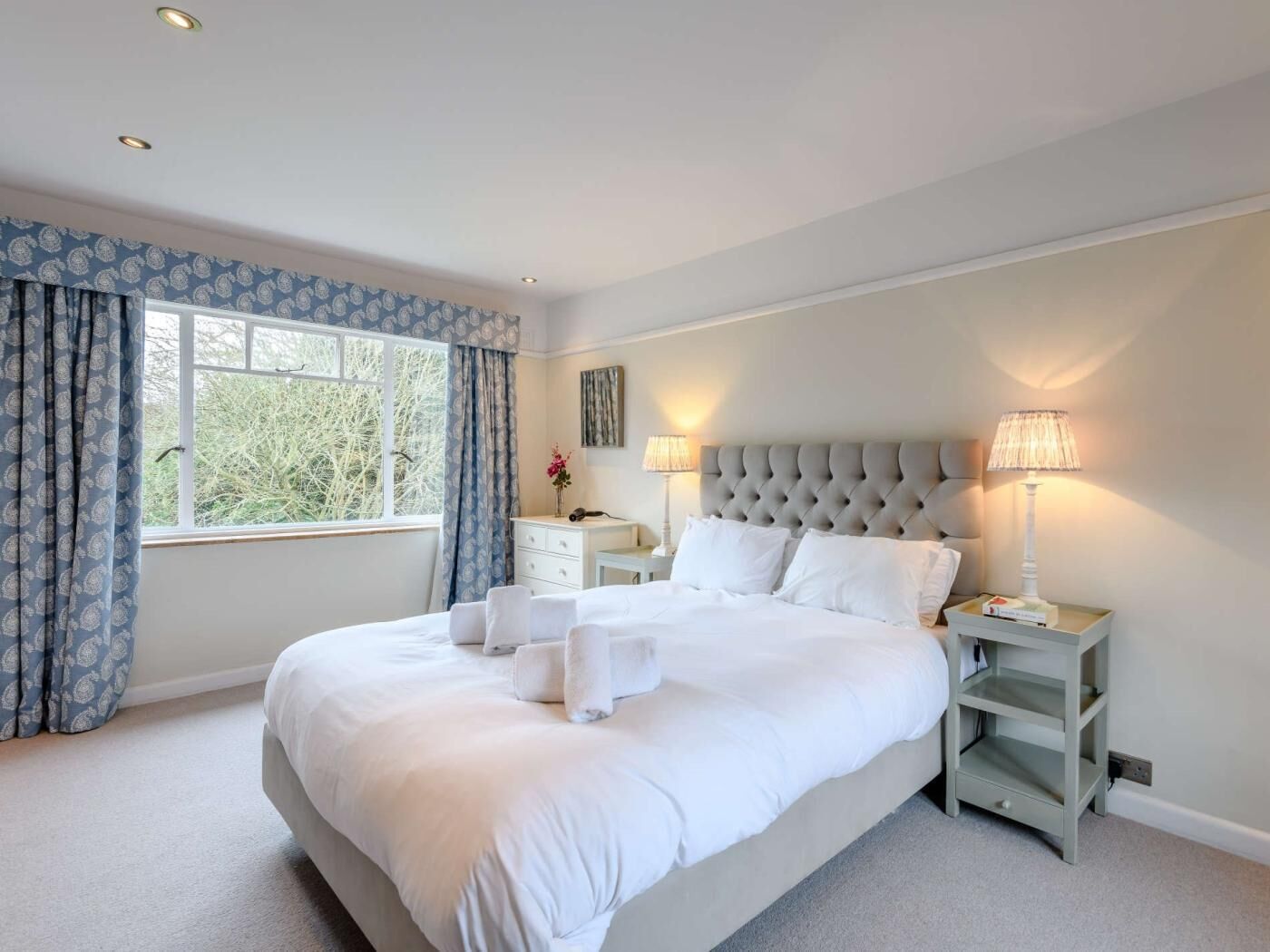 A bedroom with a large bed, two bedside tables, a nightstand, a window with curtains, and a lamp.