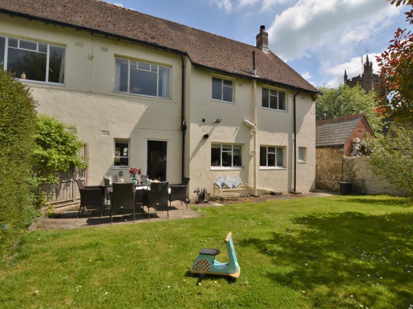 Une maison à deux étages avec un jardin, une terrasse dotée de mobilier d’extérieur et un petit scooter bleu.