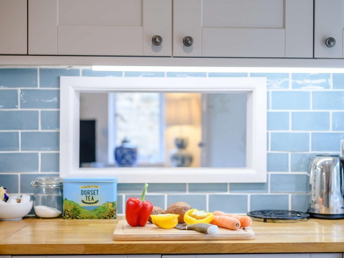 Una cocina con ventana, tabla de cortar con verduras, una caja de té Dorset y una tetera.