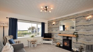 A living room with a fireplace, a television, a sofa, and a large window. - 2 Bed in Moelfre (oc-75013) (Moelfre)