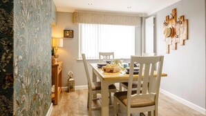 A dining area with a table set for four, a window with blinds, a wall-mounted hat rack, and a lamp on a side table. - 2 Bed in Moelfre (oc-75013) (Moelfre)
